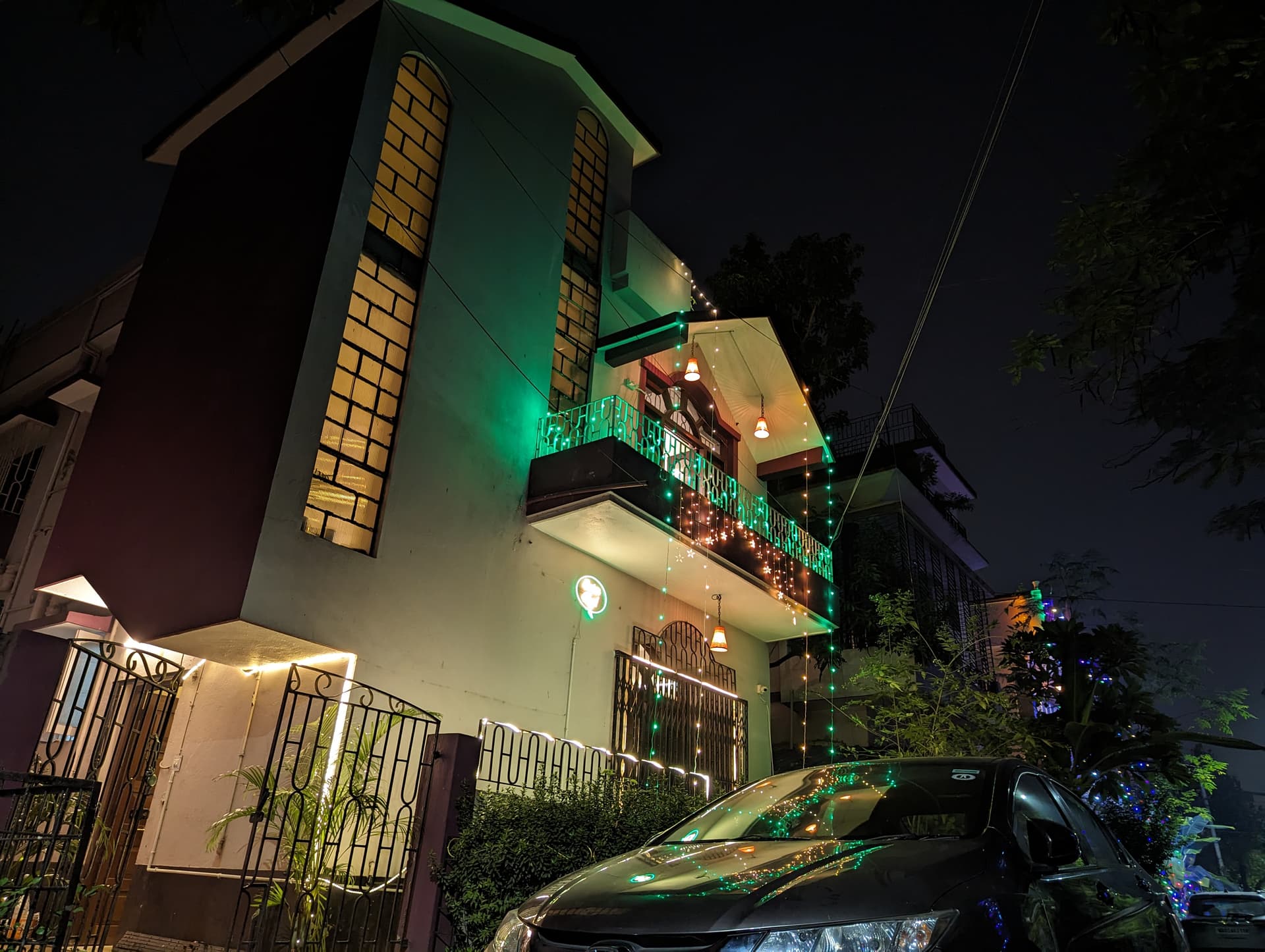 Wide evening photograph of Guten Tag Kolkata bungalow with illuminated arched window column, lit balconies, and green property sign
